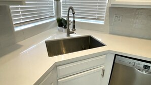 Una cocina con un lavaplatos de acero inoxidable, armarios blancos, un lavavajillas Bosch y una ventana con persianas.