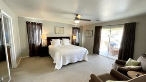 Una habitación de hotel con una cama grande, dos mesitas de noche con lámparas, un ventilador de techo, un ventanal corredero a un patio y un sillón marrón.
