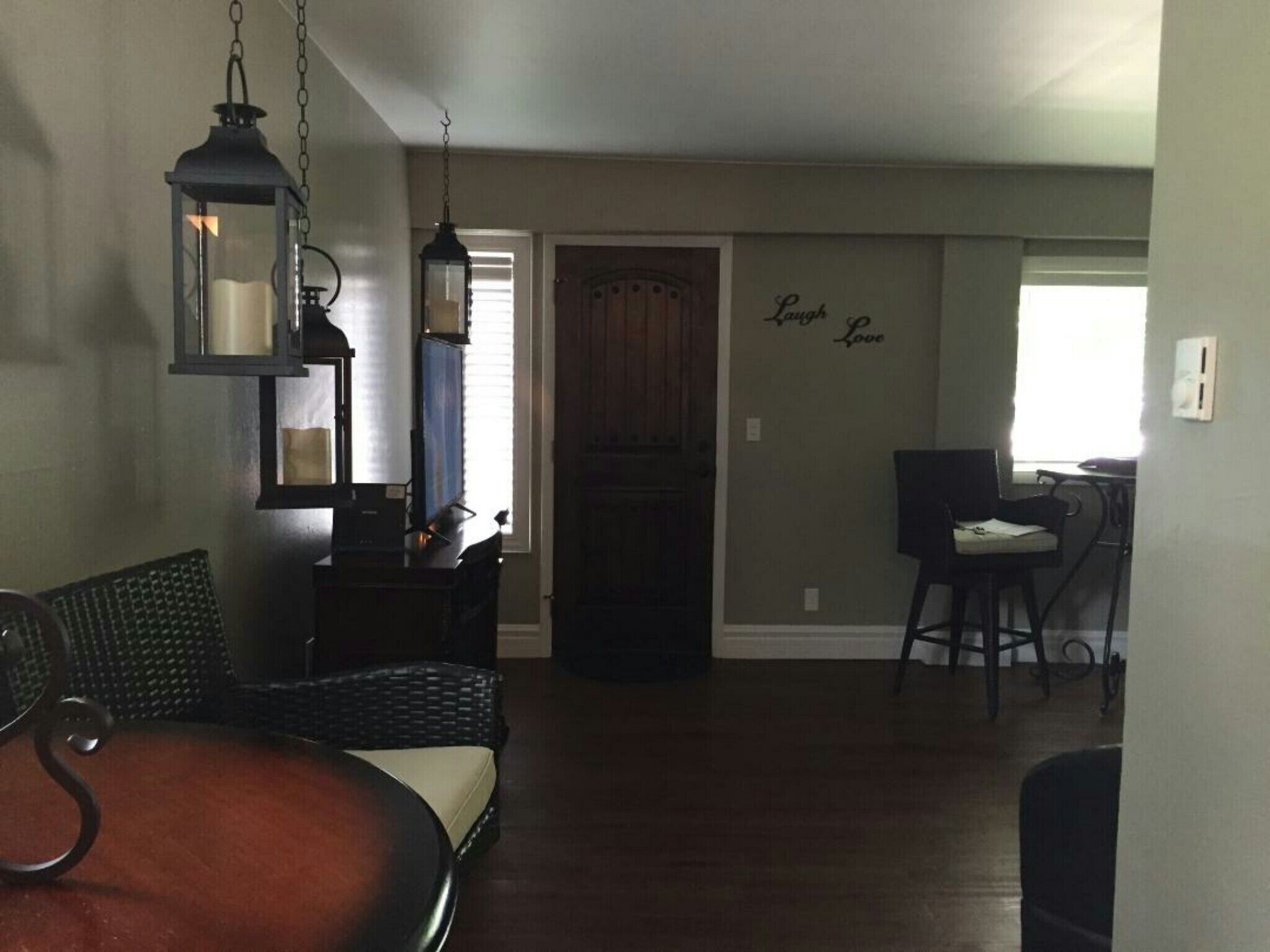 A dining area with a round table, two chairs, and a hanging lantern. There is a wooden door and a wall with a 'Laugh & Love' sign.