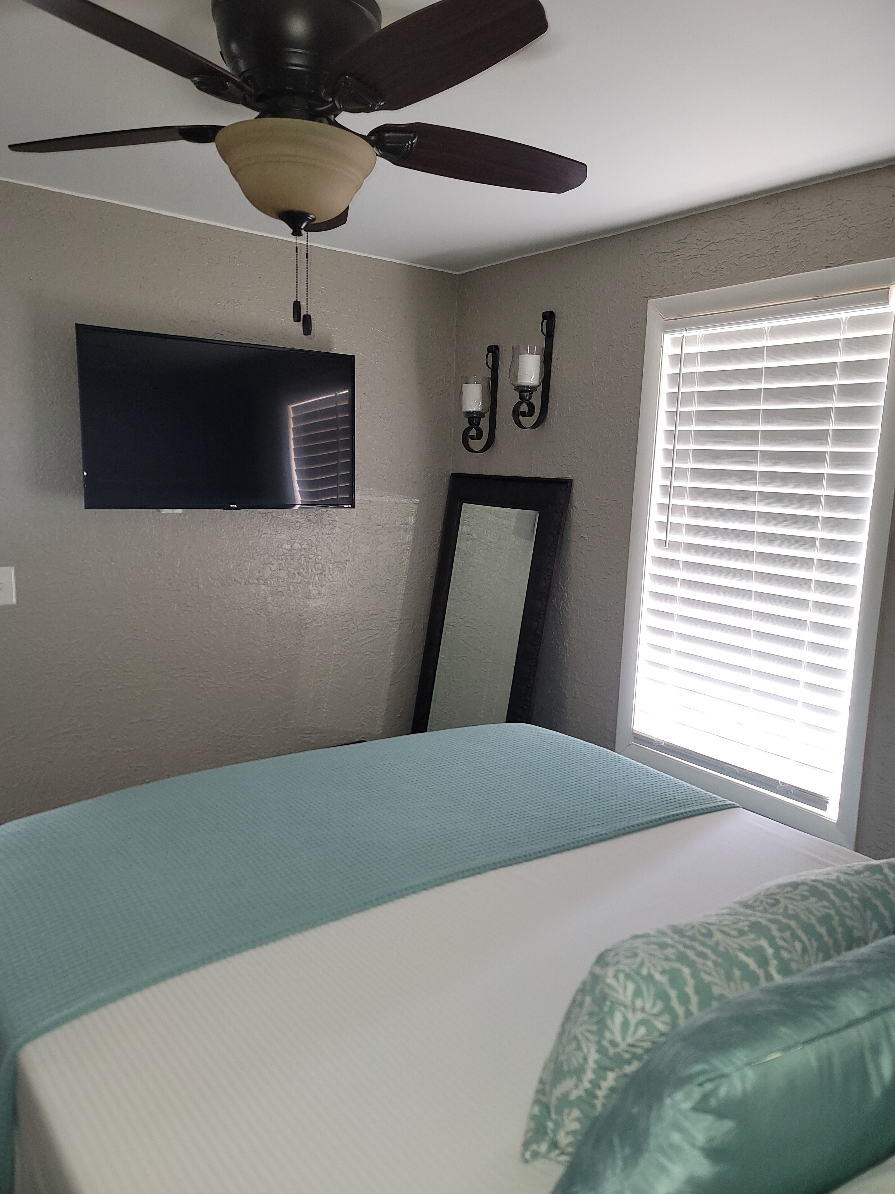 A bedroom with a bed, a ceiling fan, a flat-screen TV, a wall-mounted mirror, and a window with blinds.