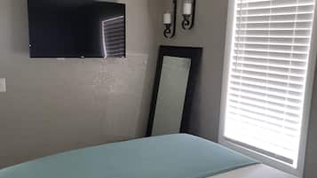A bedroom with a bed, a ceiling fan, a flat-screen TV, a wall-mounted mirror, and a window with blinds.
