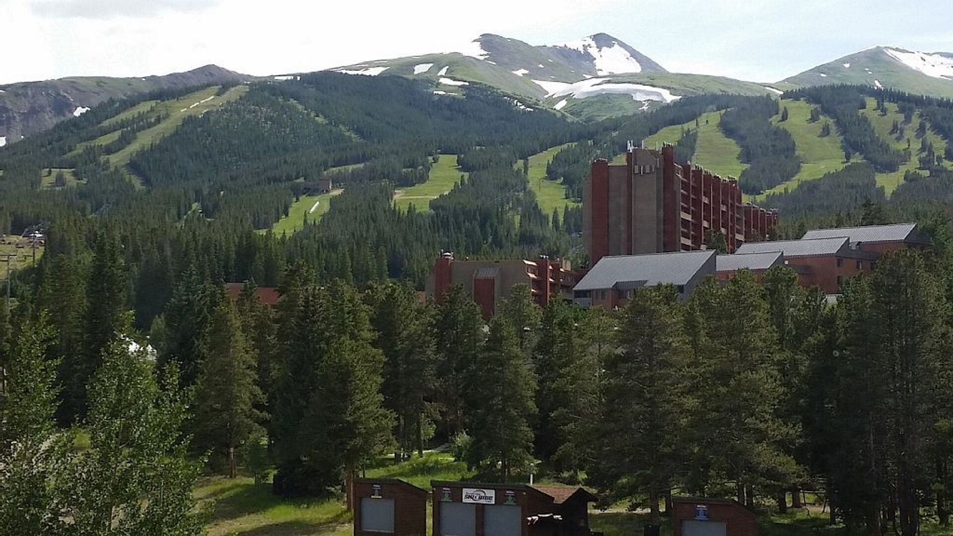 A building with multiple red brick structures and a large tower, surrounded by dense forest and snow-capped mountains.