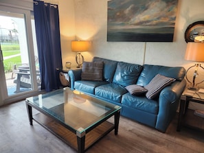 A living room with a glass coffee table, blue leather sofa, and a painting on the wall.