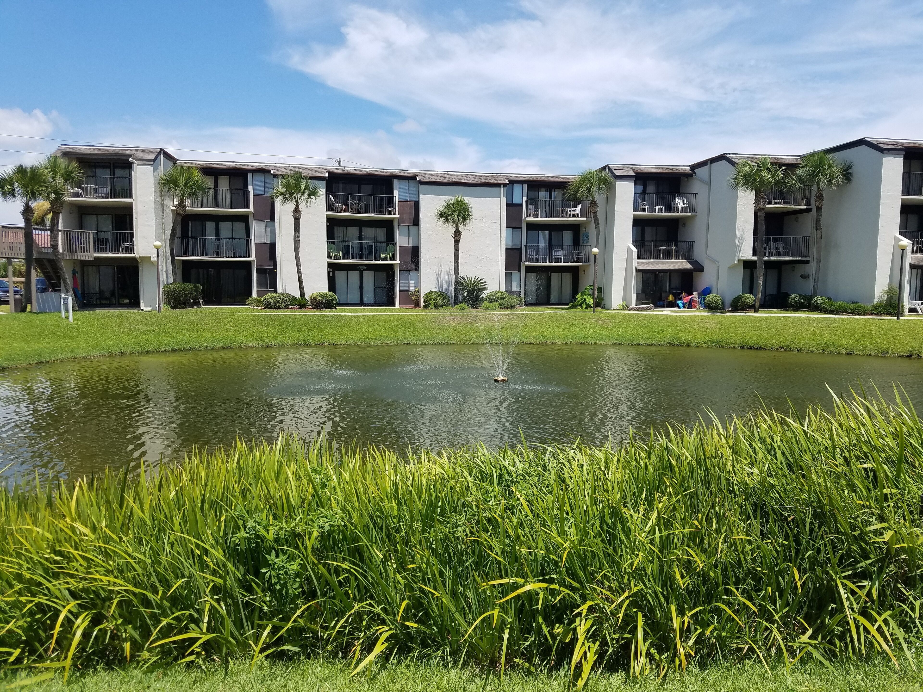 A multi-story residential building with balconies, a small pond, and lush greenery.