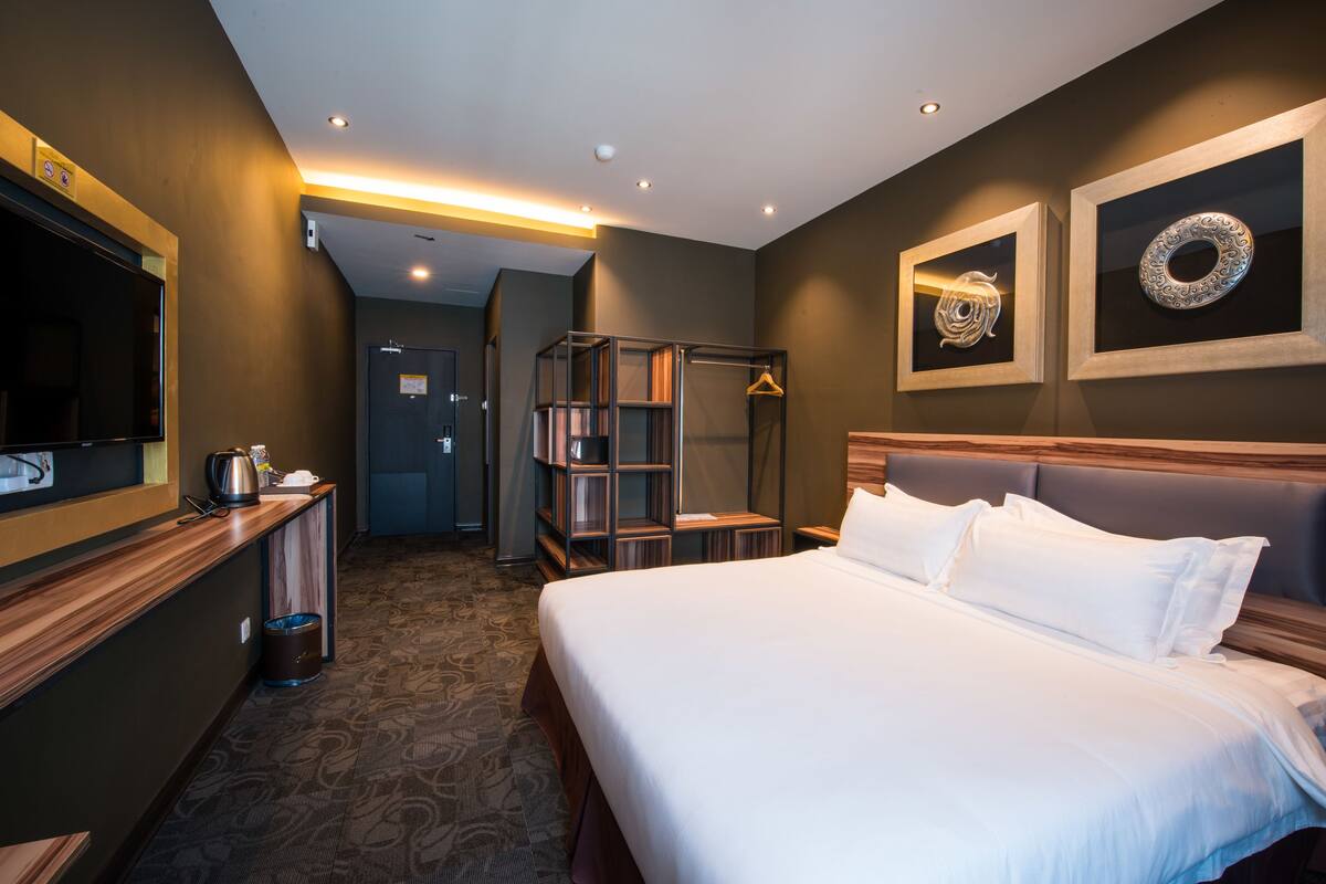 A hotel room with a large bed, a TV, a wooden desk, a bookshelf, and a framed artwork on the wall.