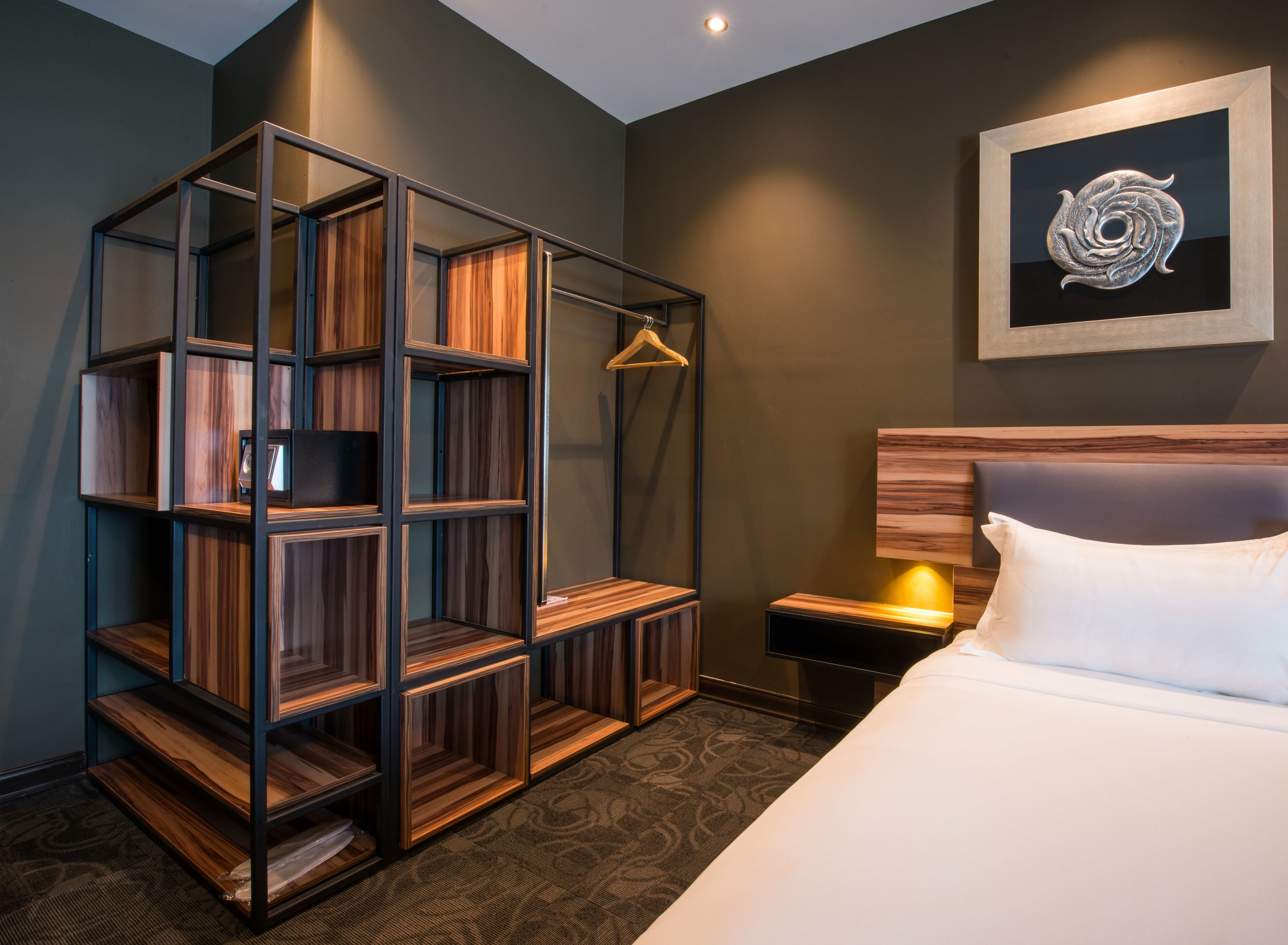 A hotel room with a bed, a wooden shelving unit, a wall-mounted artwork, and a nightstand with a lamp.
