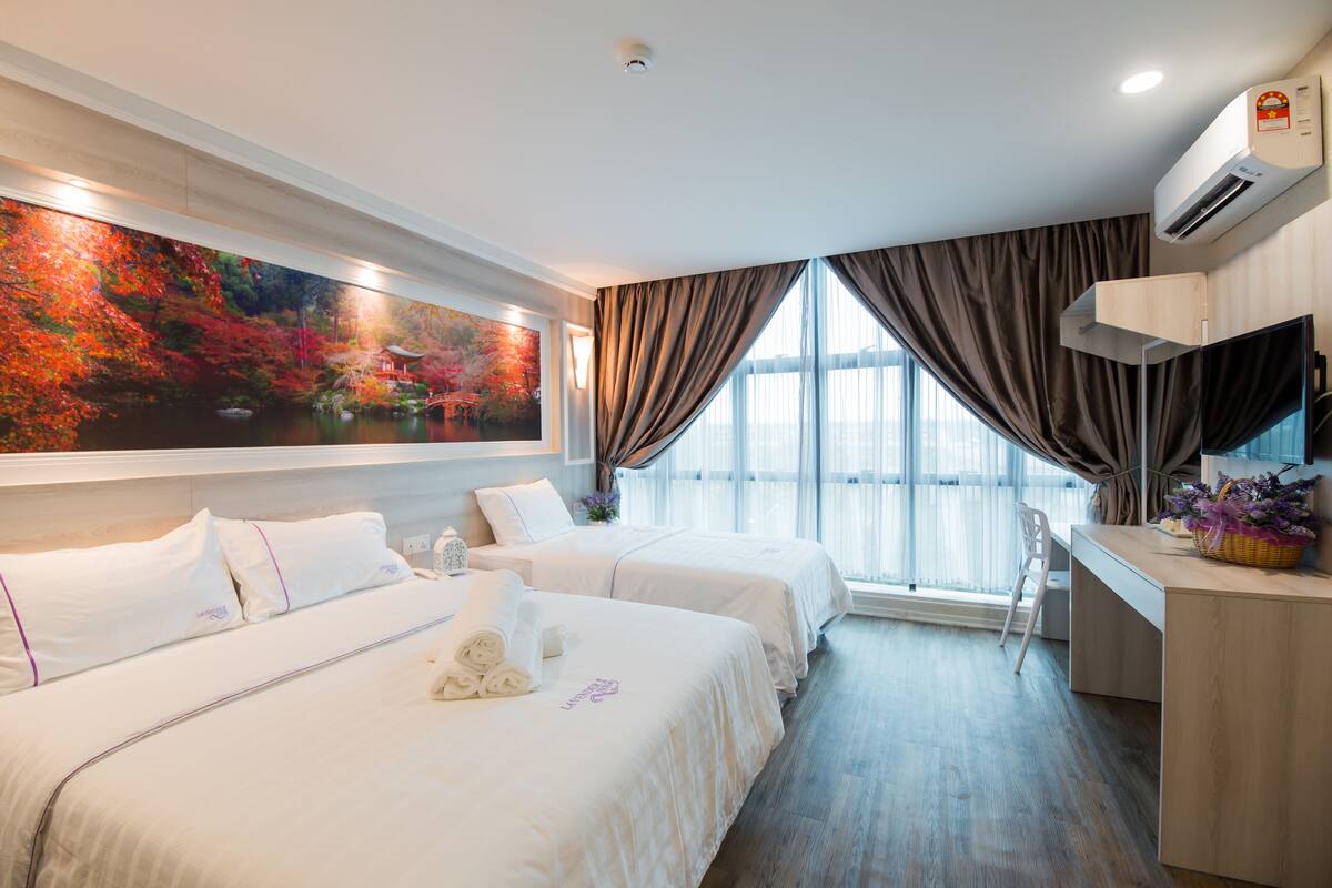 A hotel room with two beds, a desk, a large window with curtains, an air conditioning unit, and a painting of a landscape.