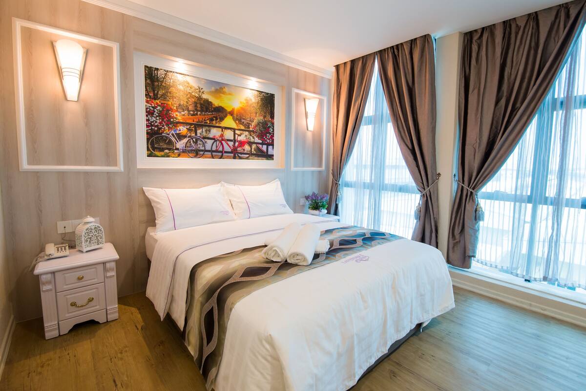 A well-lit hotel room with a large bed, a nightstand with a lamp, and a painting above the bed.
