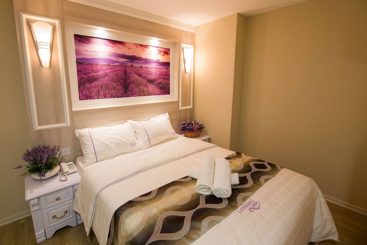A neatly made bed with a floral-patterned bedspread, a bedside lamp, a nightstand with a telephone and a vase of flowers, and a large framed picture above the bed.