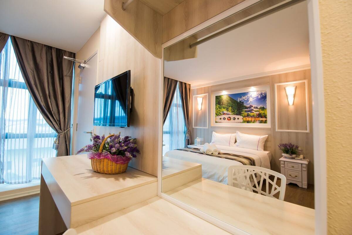 A hotel room with a large mirror, a bed, a bedside table with a basket of flowers, a TV, and a painting on the wall.