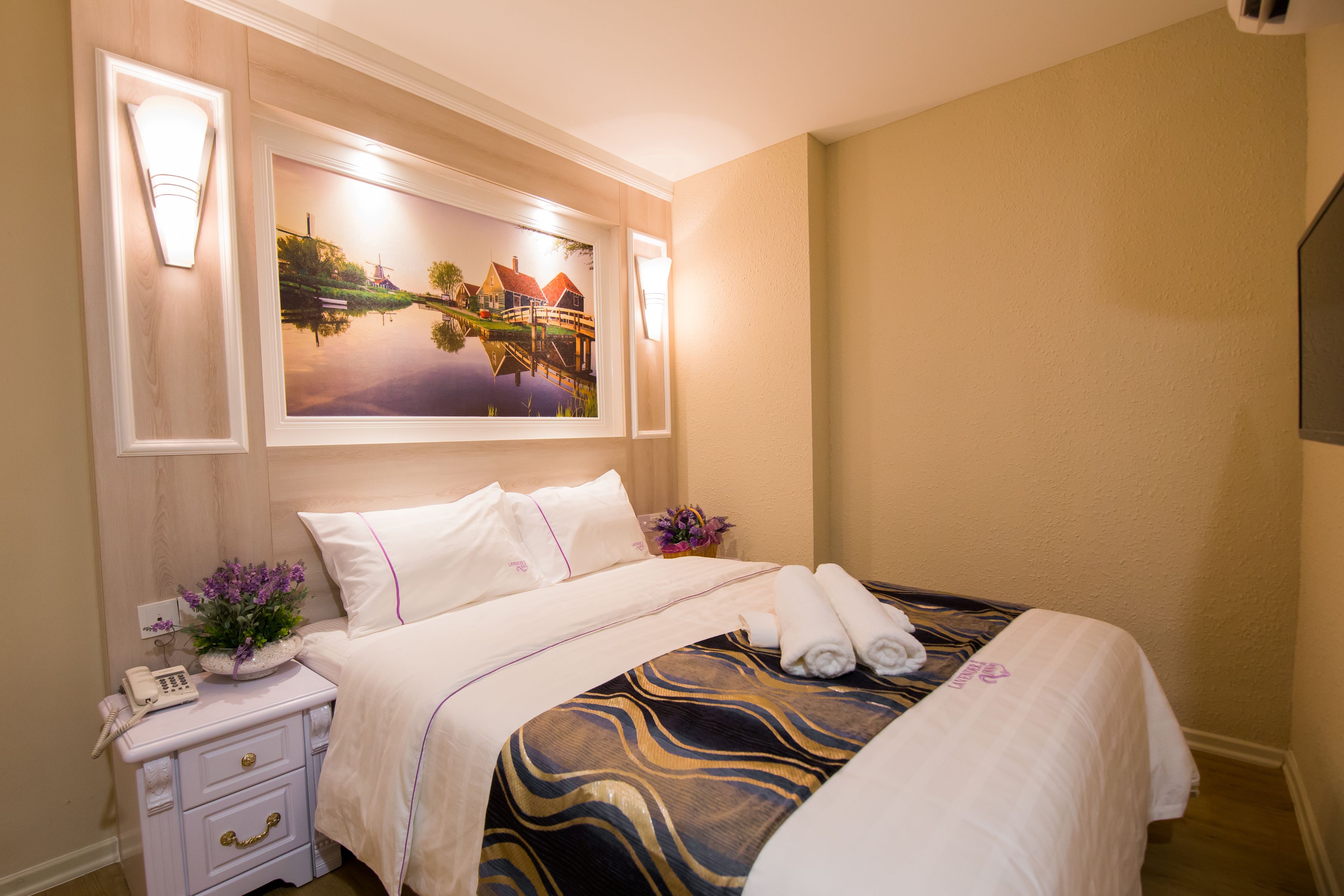 Un lit bien fait, recouvert d’une couvre-lit à motifs, une table de chevet avec une lampe et un téléphone, et un tableau accroché au-dessus du lit.