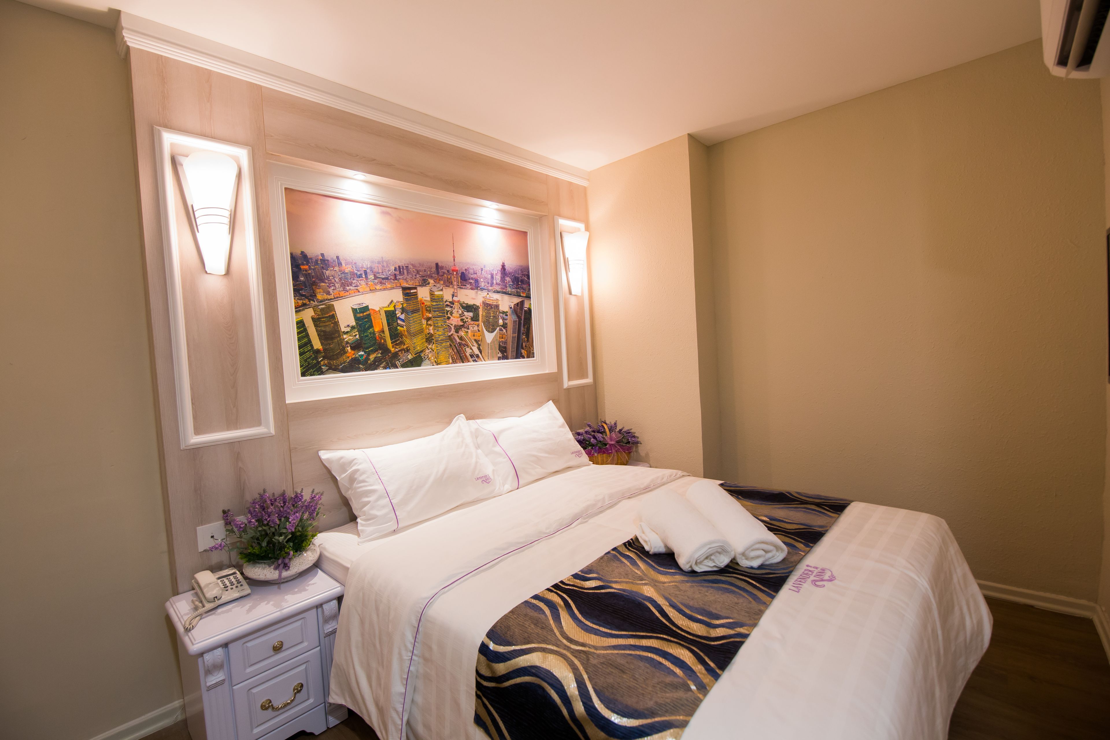 A neatly made bed with a patterned bedspread, a bedside table with a lamp and a phone, and a wall-mounted artwork depicting a cityscape.