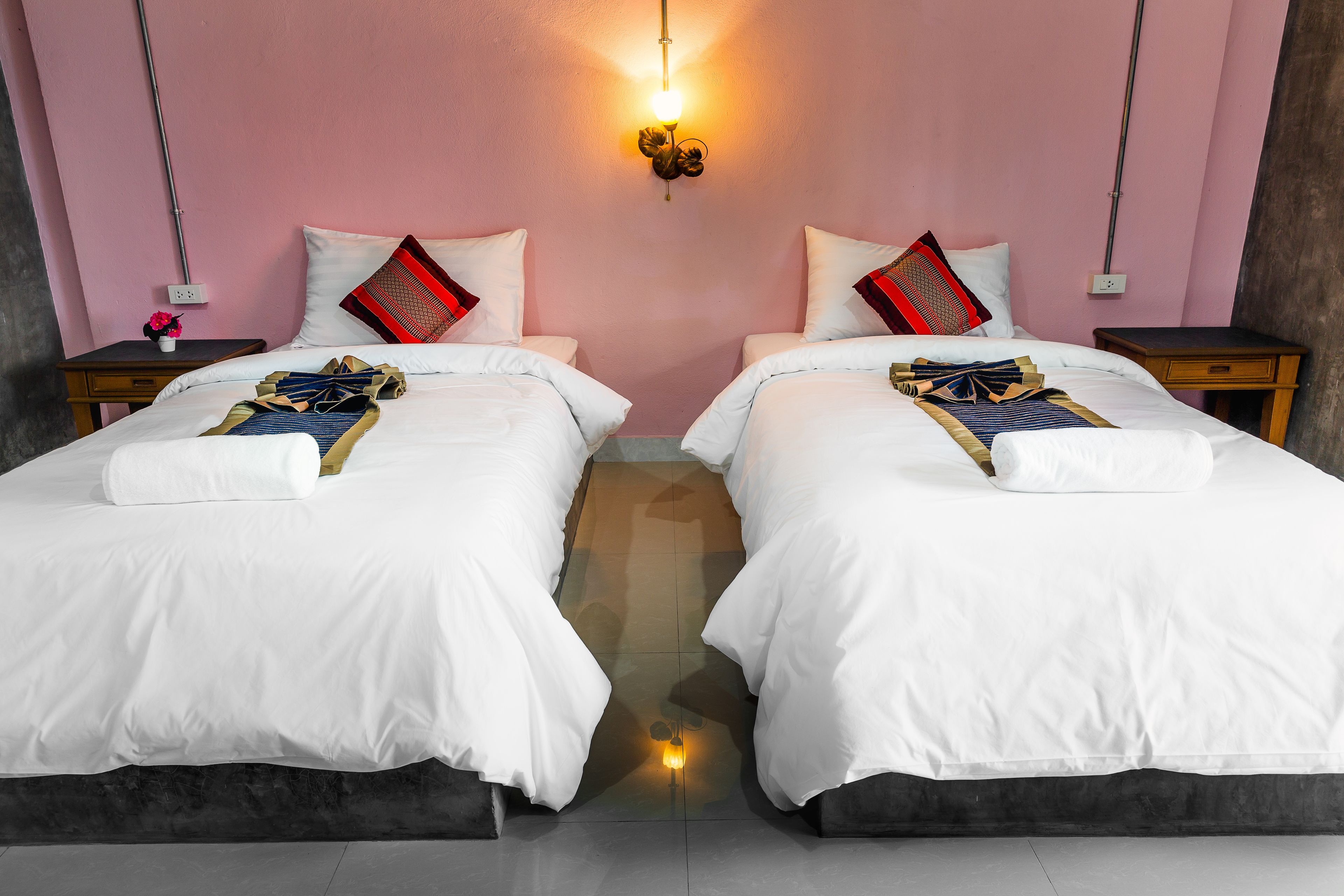 A hotel room with two beds, each with white bedding and red and black pillows. There is a wall-mounted light fixture above the beds, and a small table with a vase of flowers on one side.
