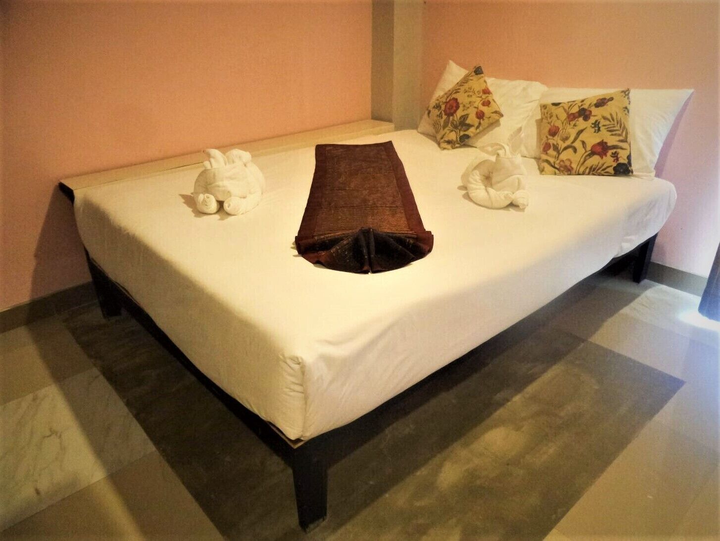 Una cama bien tendida con una manta marrón, almohadas blancas y una almohada estampada sobre un piso a cuadros.