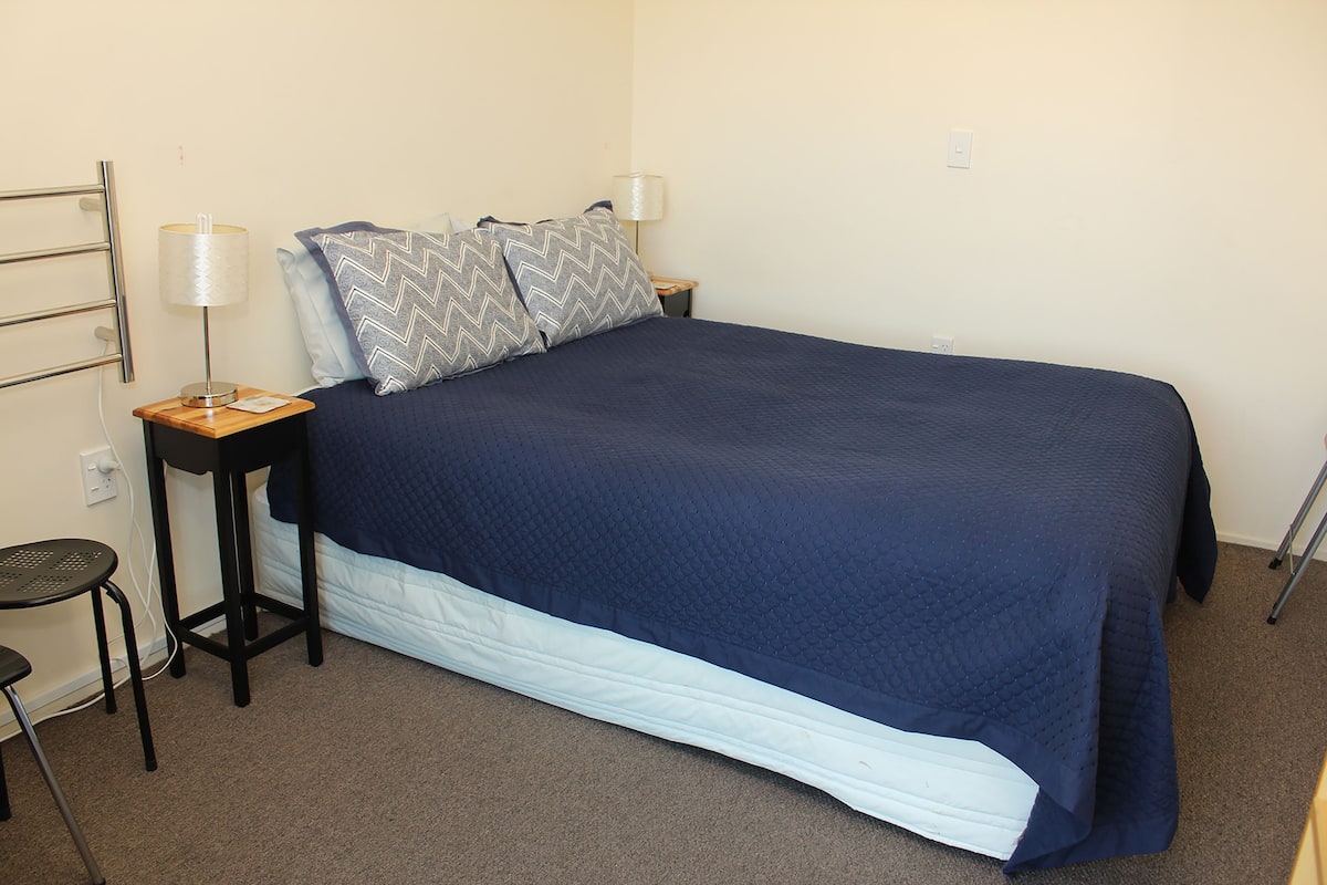 A neatly made bed with a blue cover, two bedside tables with lamps, and a chair.