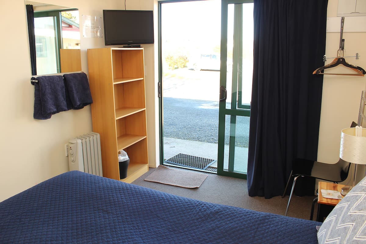 A hotel room with a bed, a wooden wardrobe, a television, a window with curtains, and a door leading outside.