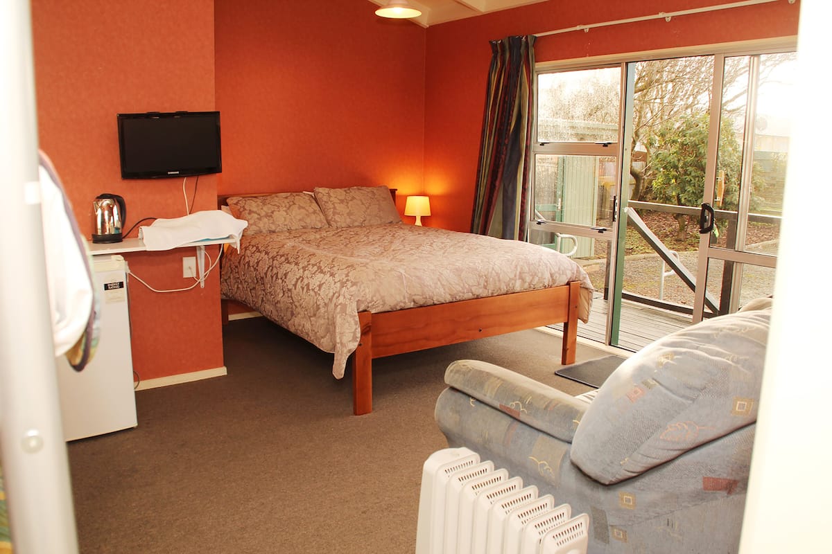 A bedroom with a bed, a bedside table with a lamp, a television on the wall, and a window with curtains.