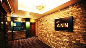A hotel lobby with a brick wall, a flat-screen TV displaying "THE ANN", and a check-in counter. - Hotel The Ann (Changwon)