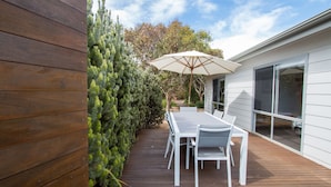 Une terrasse en bois avec une table et des chaises blanches, un parasol et une haie bien entretenue.