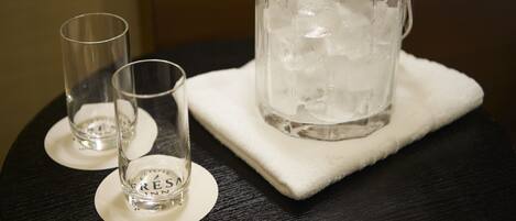 A clear glass pitcher with ice and a spoon on a white cloth, accompanied by two empty glasses on a dark table.