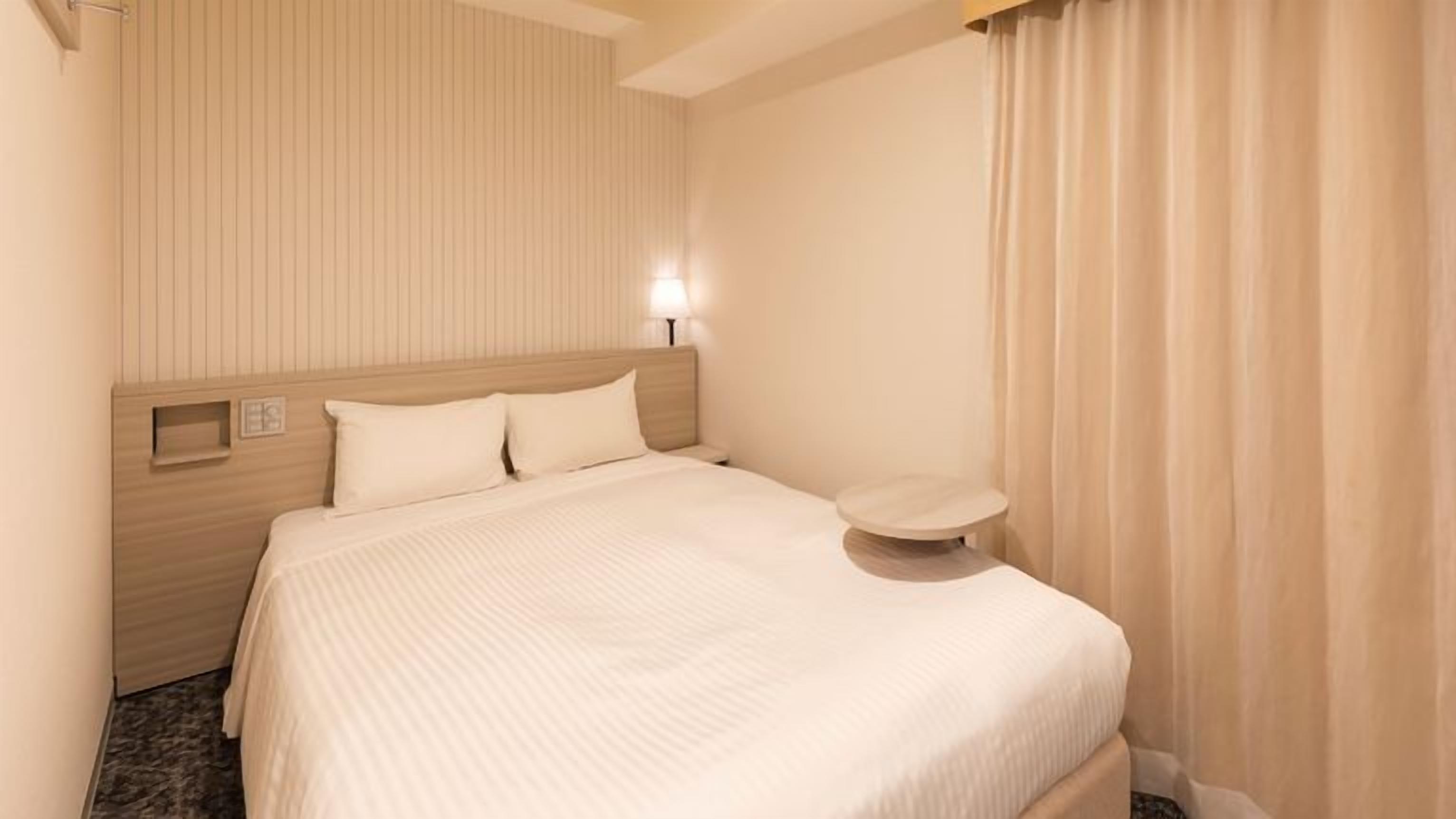 Habitación de hotel con cama, mesita de noche, lámpara de pared y ventana con cortinas.