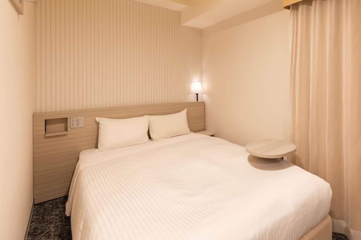 Habitación de hotel con cama, mesita de noche, lámpara de pared y ventana con cortinas.