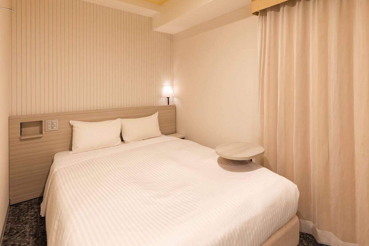 Ein Hotelzimmer mit Bett, Nachttisch, Lampe und einem Fenster mit Vorhängen.