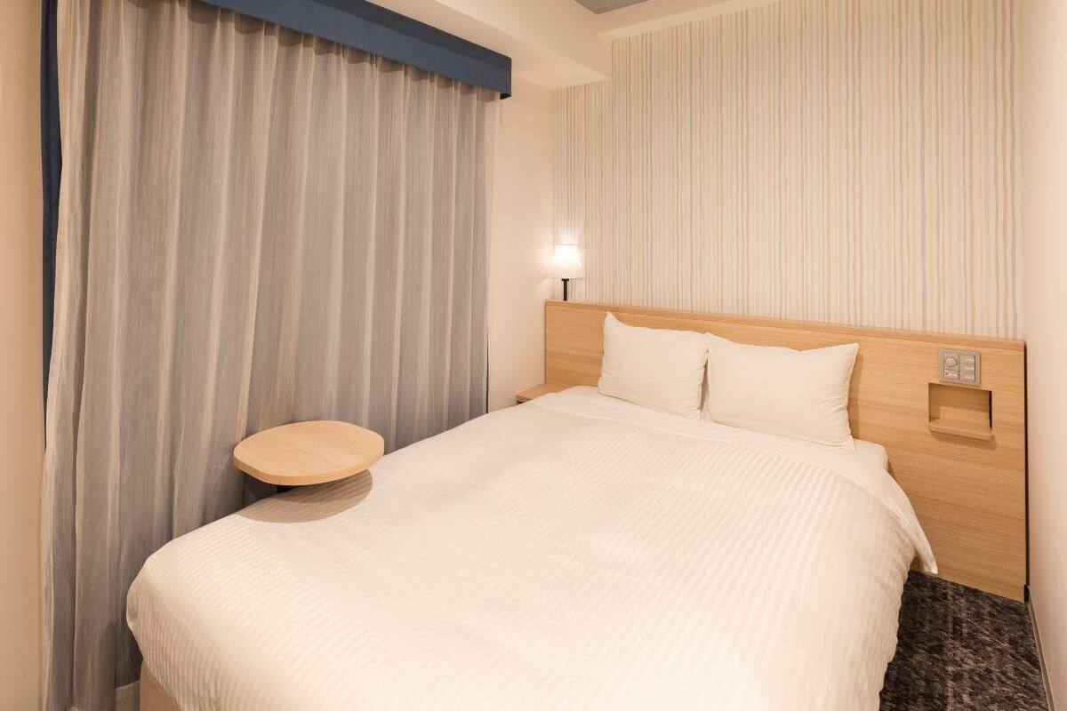 Ein Hotelzimmer mit Bett, Nachttisch, Lampe und Vorhang.