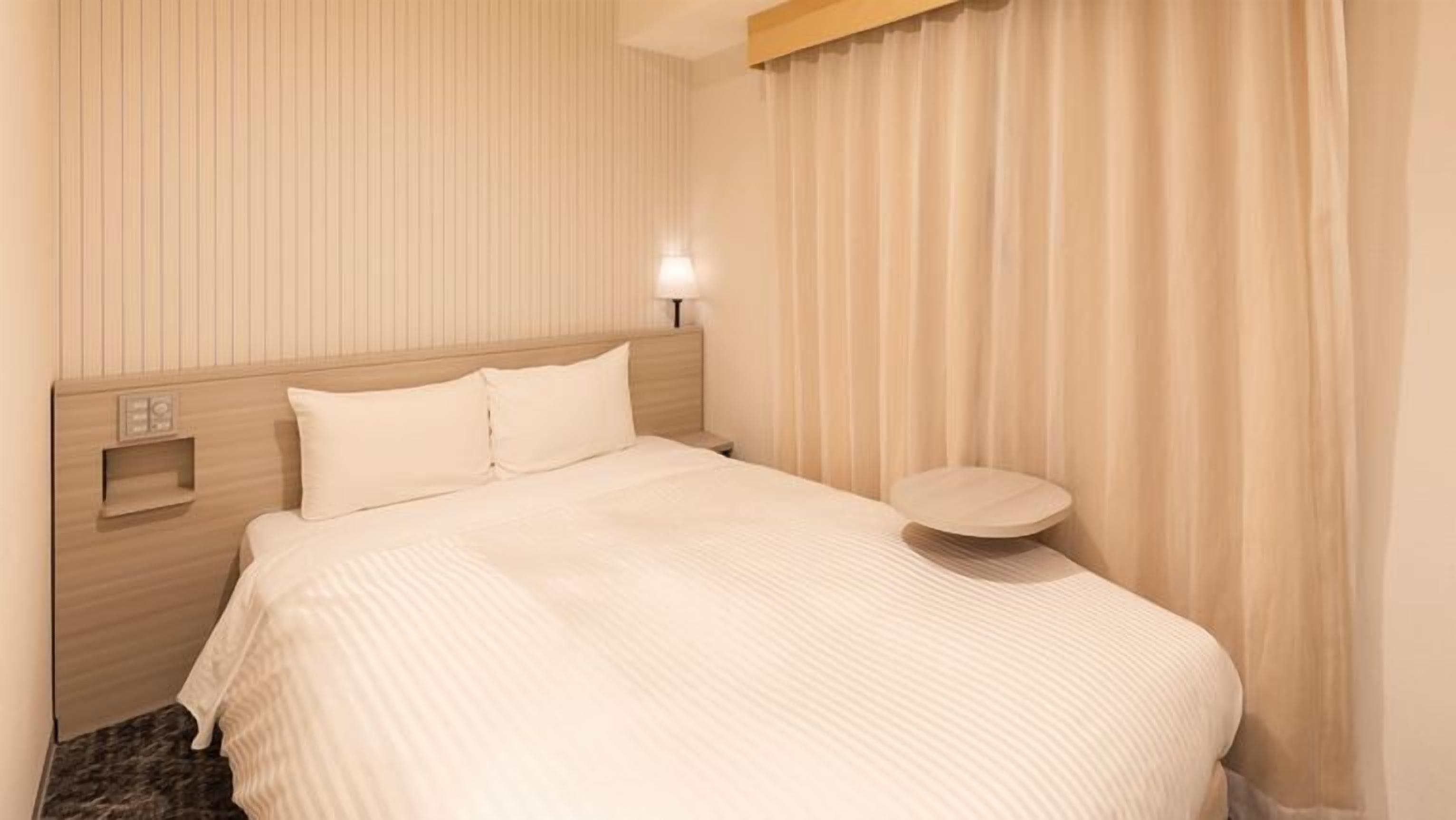 Una habitación de hotel con cama, mesita de noche, luz empotrada en la pared y ventana con cortinas.