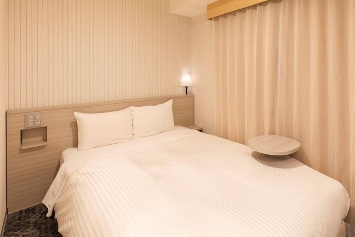 Ein Hotelzimmer mit Bett, Nachttisch, Wandleuchte und einem Fenster mit Vorhängen.