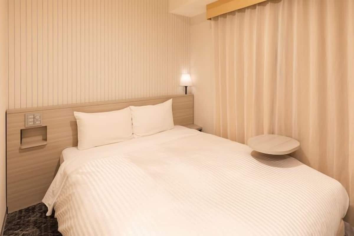 Una habitación de hotel con cama, mesita de noche, luz empotrada en la pared y ventana con cortinas.