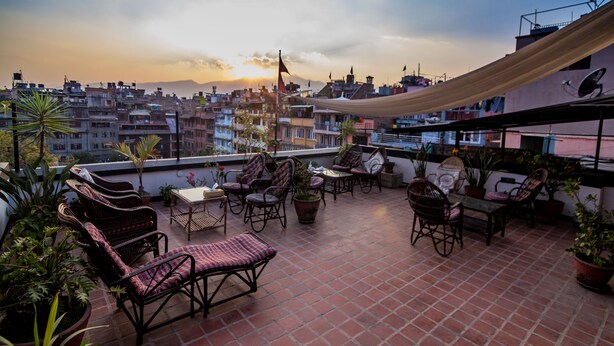 Terraza en la azotea con mobiliario de mimbre, plantas en macetas y vistas a un paisaje urbano al atardecer.
