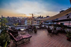 A rooftop terrace with wicker furniture, potted plants, and a view of a cityscape at sunset. - Lumbini Heritage Home (Lalitpur)