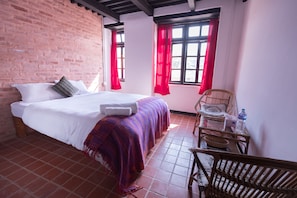 A room with a bed, a chair, a table, and a window with red curtains. - Lumbini Heritage Home (Lalitpur)