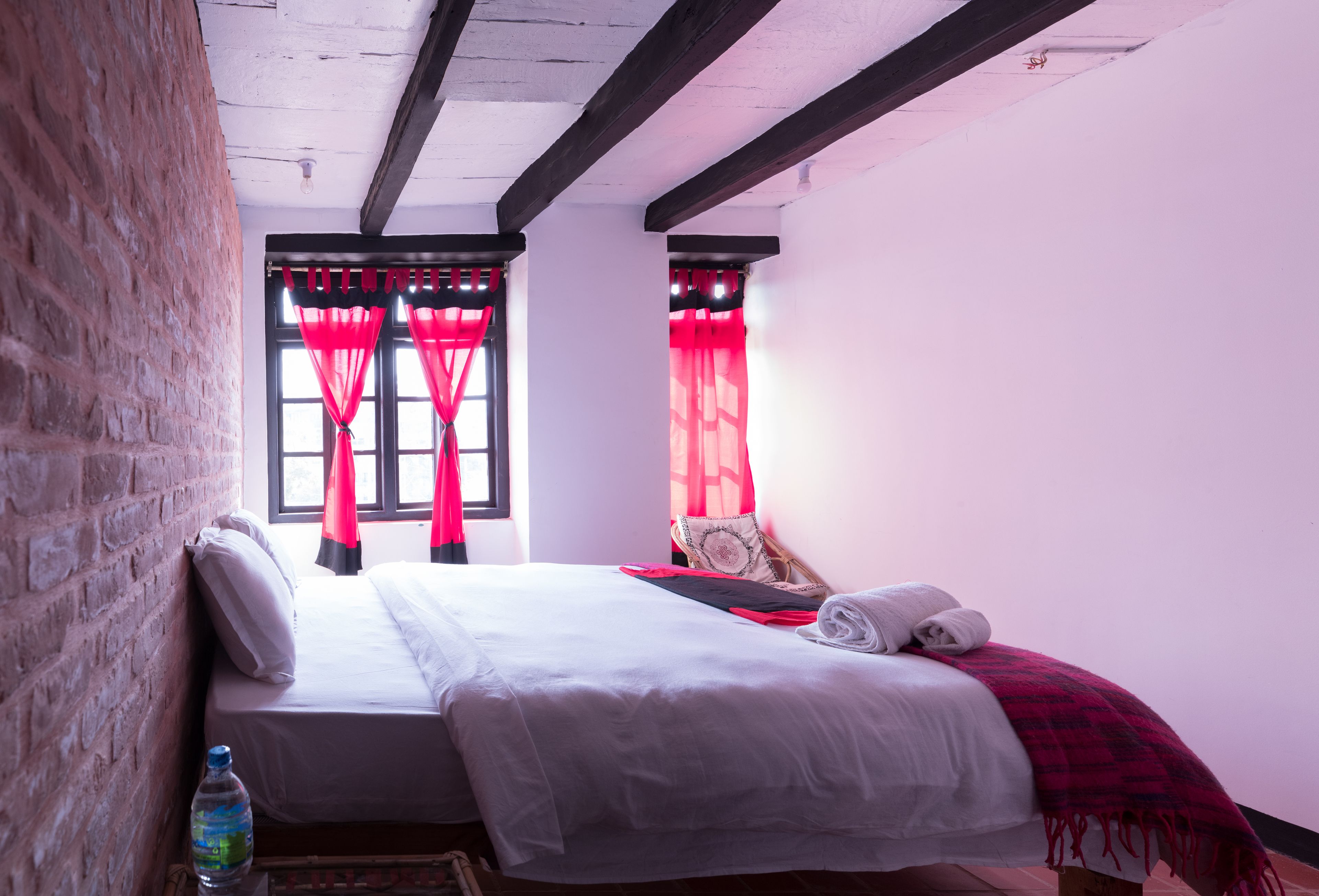 A neatly made bed with white linens, a red and black checkered blanket, and a rolled towel. There is a bottle of water on the floor beside the bed.