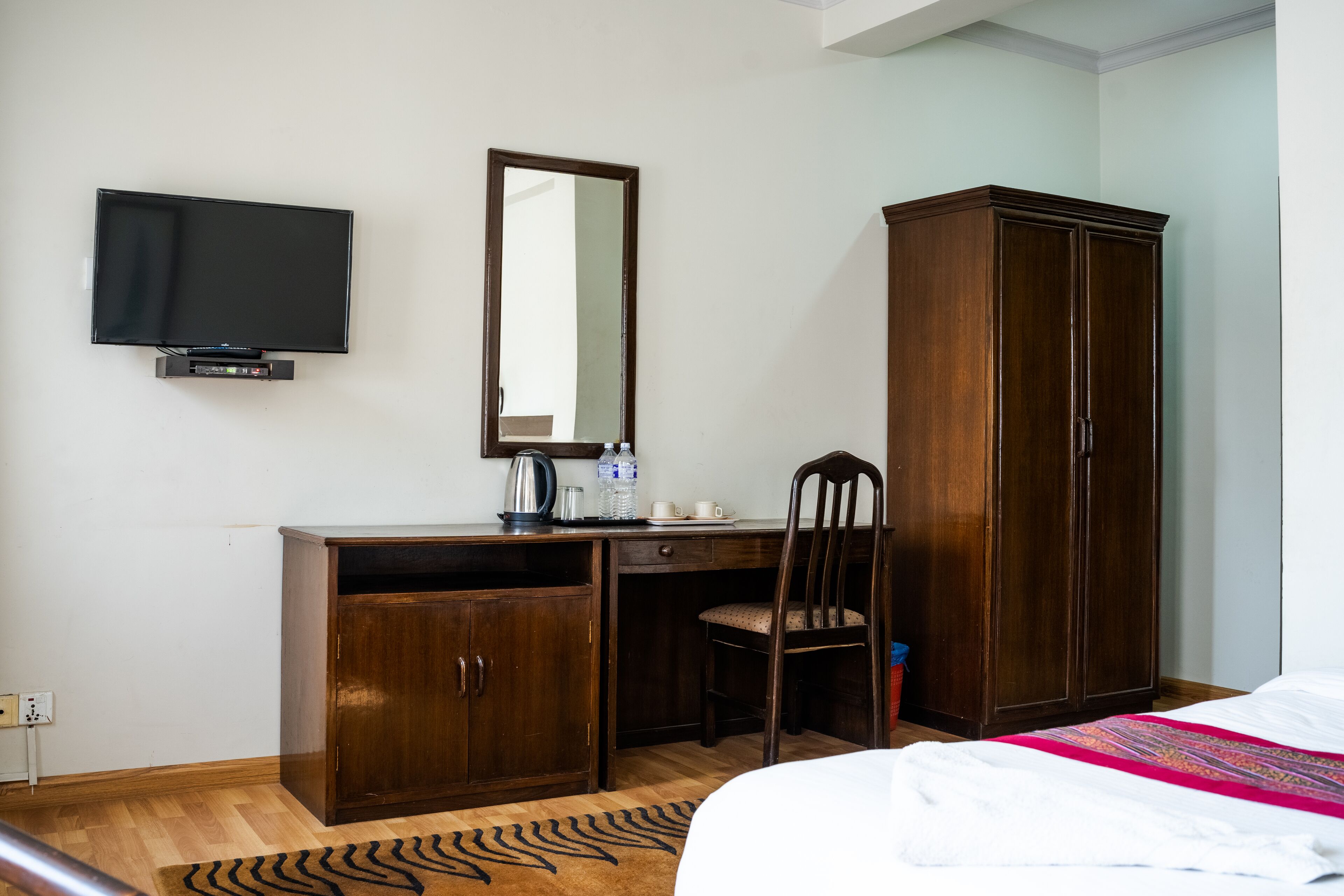 A bedroom with a bed, a wooden wardrobe, a desk with a chair, a television, and a mirror.