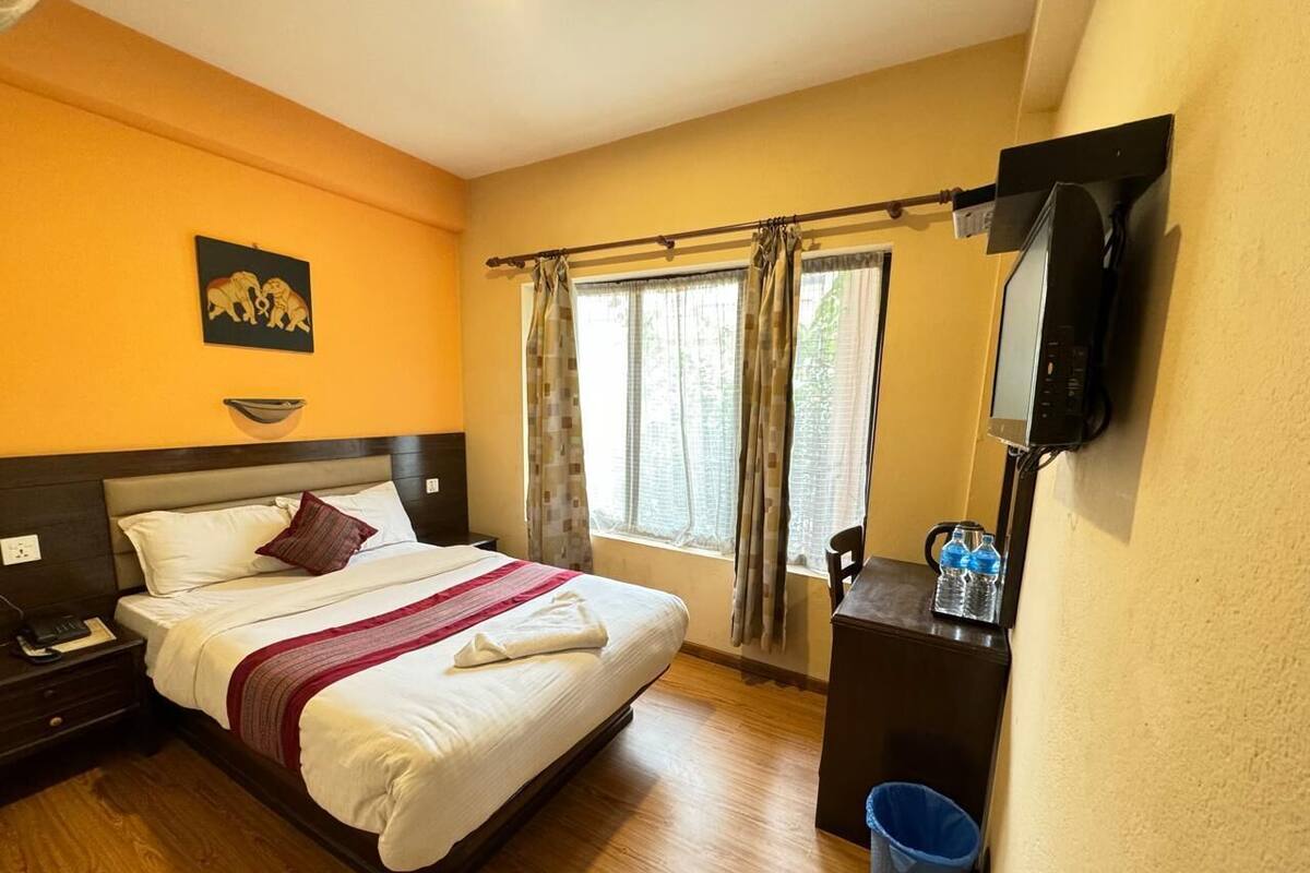 A hotel room with a bed, bedside table, a desk with a chair, a TV mounted on the wall, a window with curtains, and a wall-mounted trash bin.