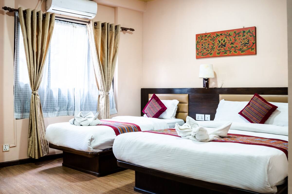 A hotel room with two beds, a window with curtains, a wall-mounted air conditioner, a nightstand with a lamp, and a decorative wall piece.