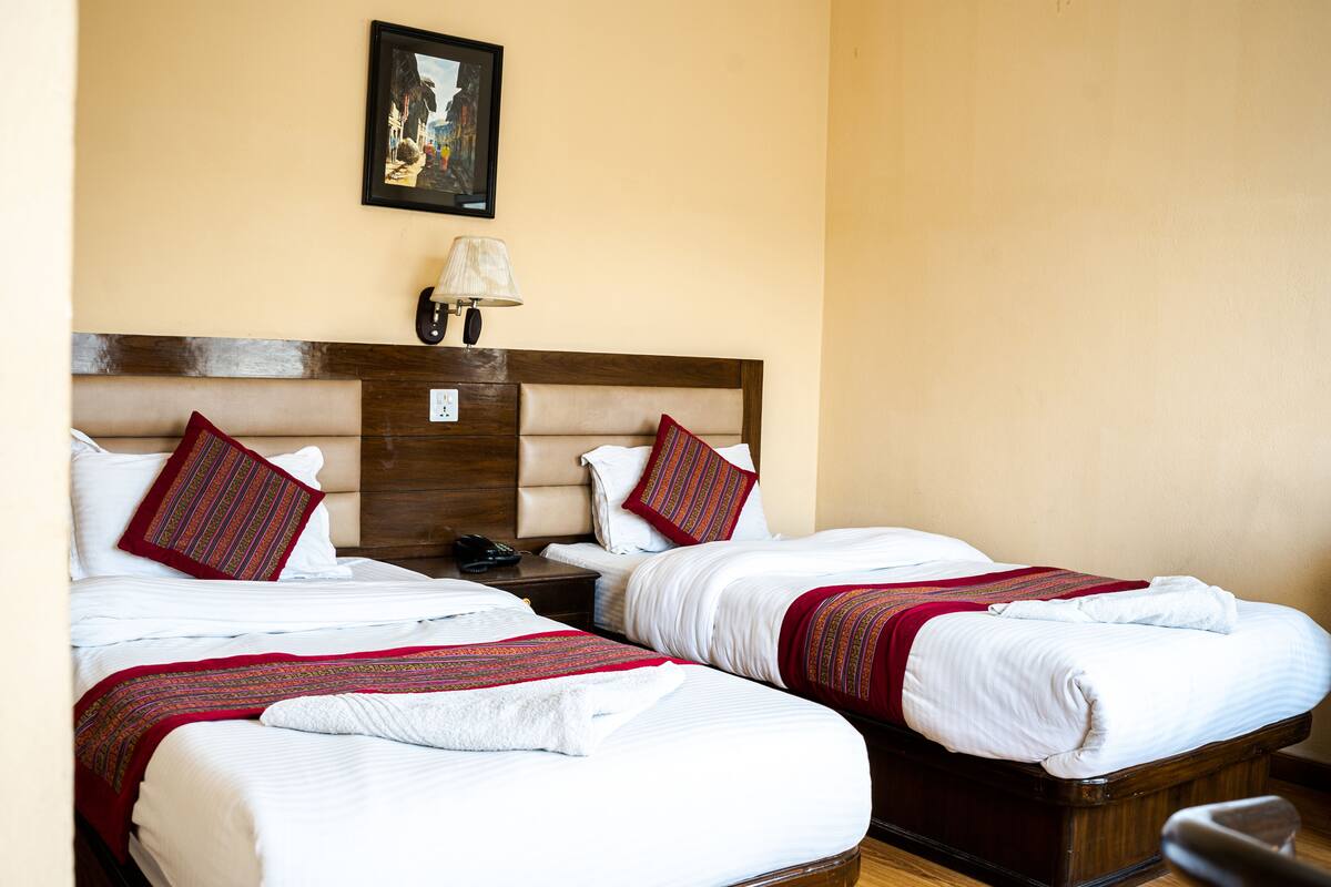 Two double beds with white linens and red accents, a wooden headboard, a nightstand with a lamp, and a framed picture on the wall.