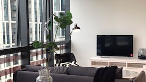 Un salon moderne avec un canapé, une table basse en verre surmontée d’un vase, un téléviseur et une grande fenêtre équipée de stores.