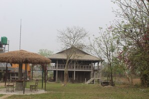 A rustic wooden house with a thatched roof, a balcony, and a staircase leading to an upper level. - Shanta Ghar Resort (Madi)