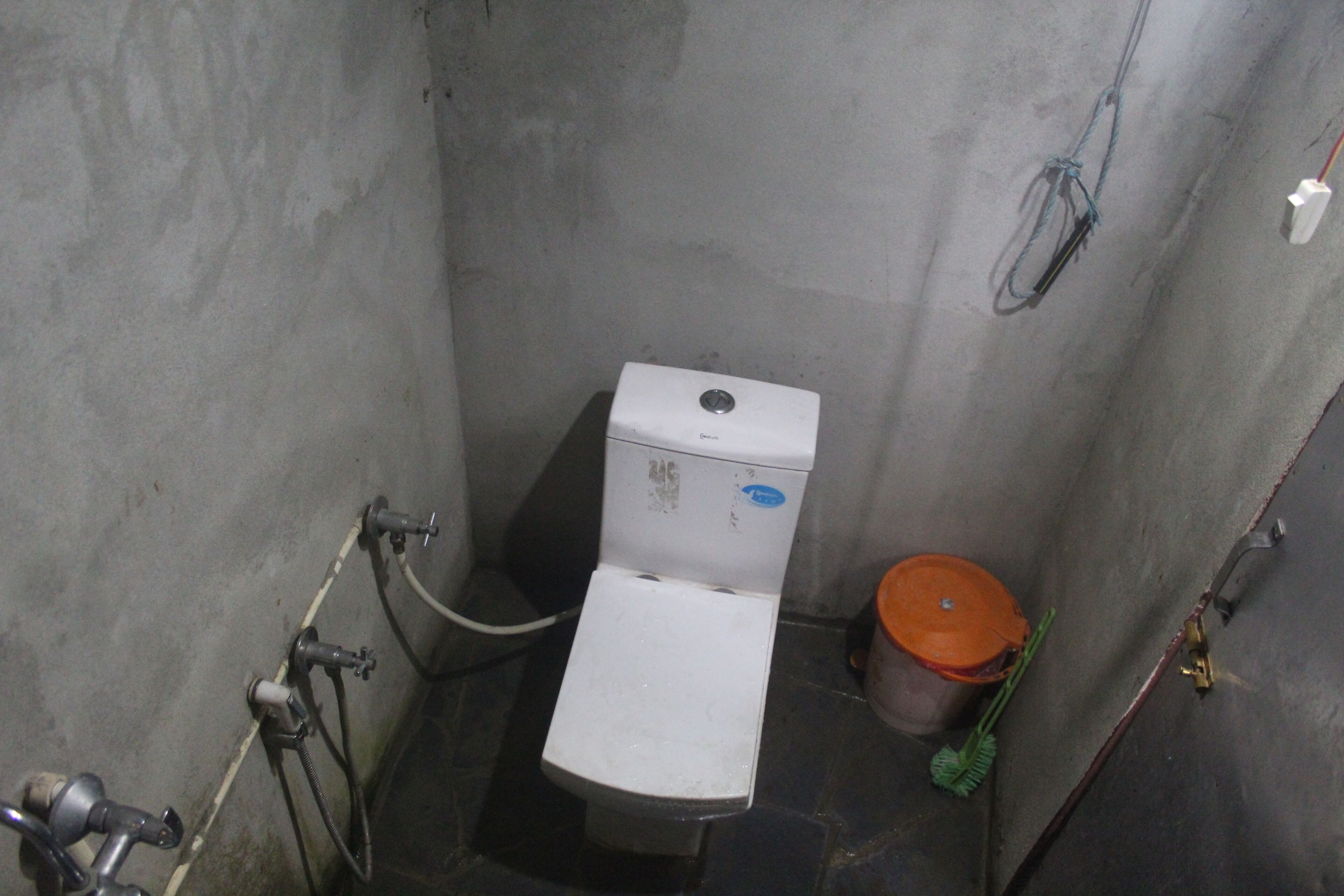 A small, enclosed bathroom with a toilet, a visible pipe, and a bucket.