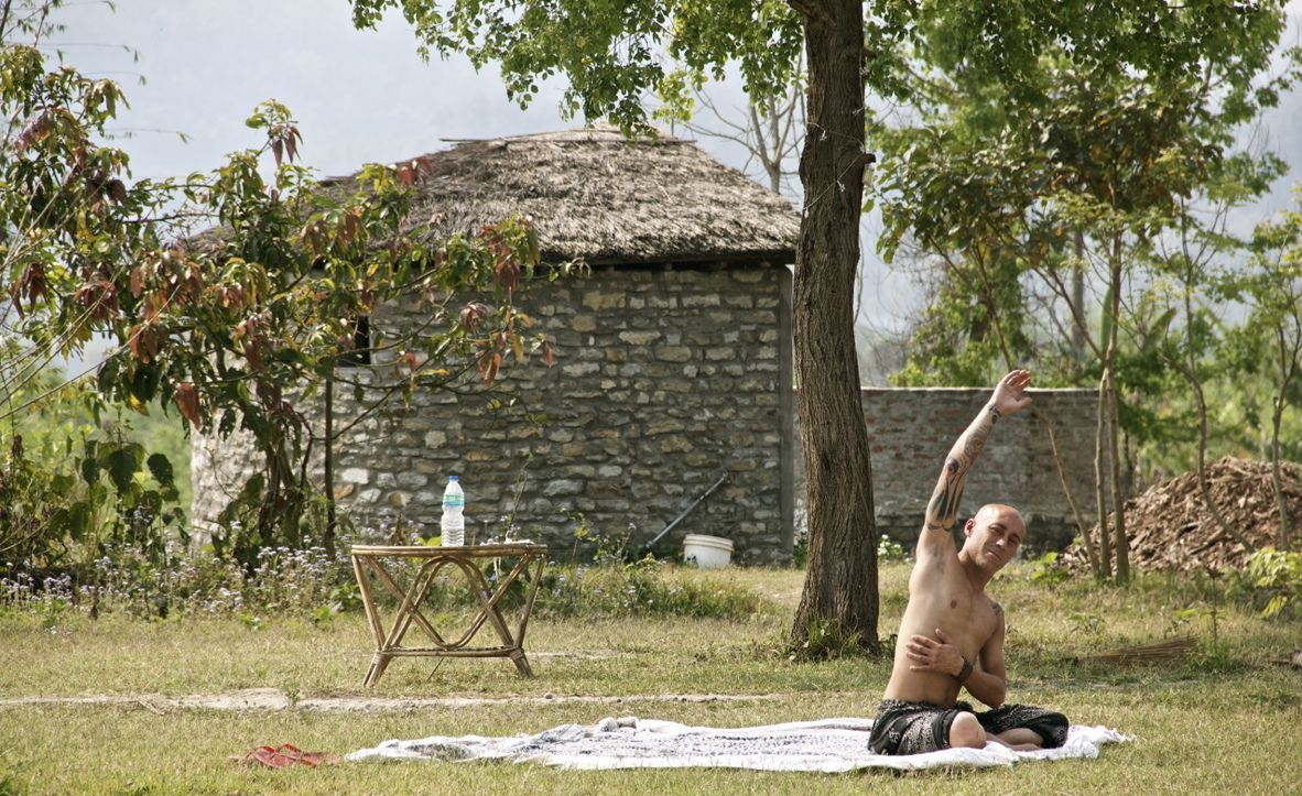Una persona sentada sobre una manta al aire libre, con un edificio de piedra y árboles de fondo.