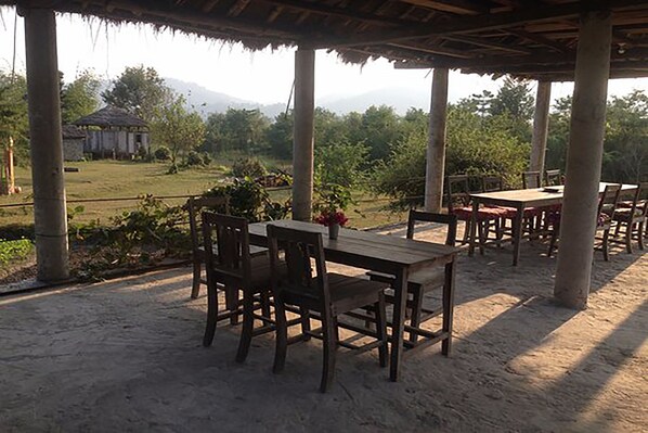 An outdoor patio with wooden tables and chairs, surrounded by columns, set against a backdrop of greenery and a distant building. - Shanta Ghar Resort (Madi)