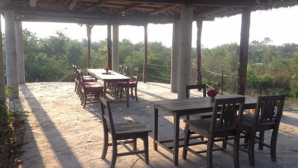 An outdoor covered patio with wooden tables and chairs, surrounded by columns, set against a backdrop of greenery. - Shanta Ghar Resort (Madi)