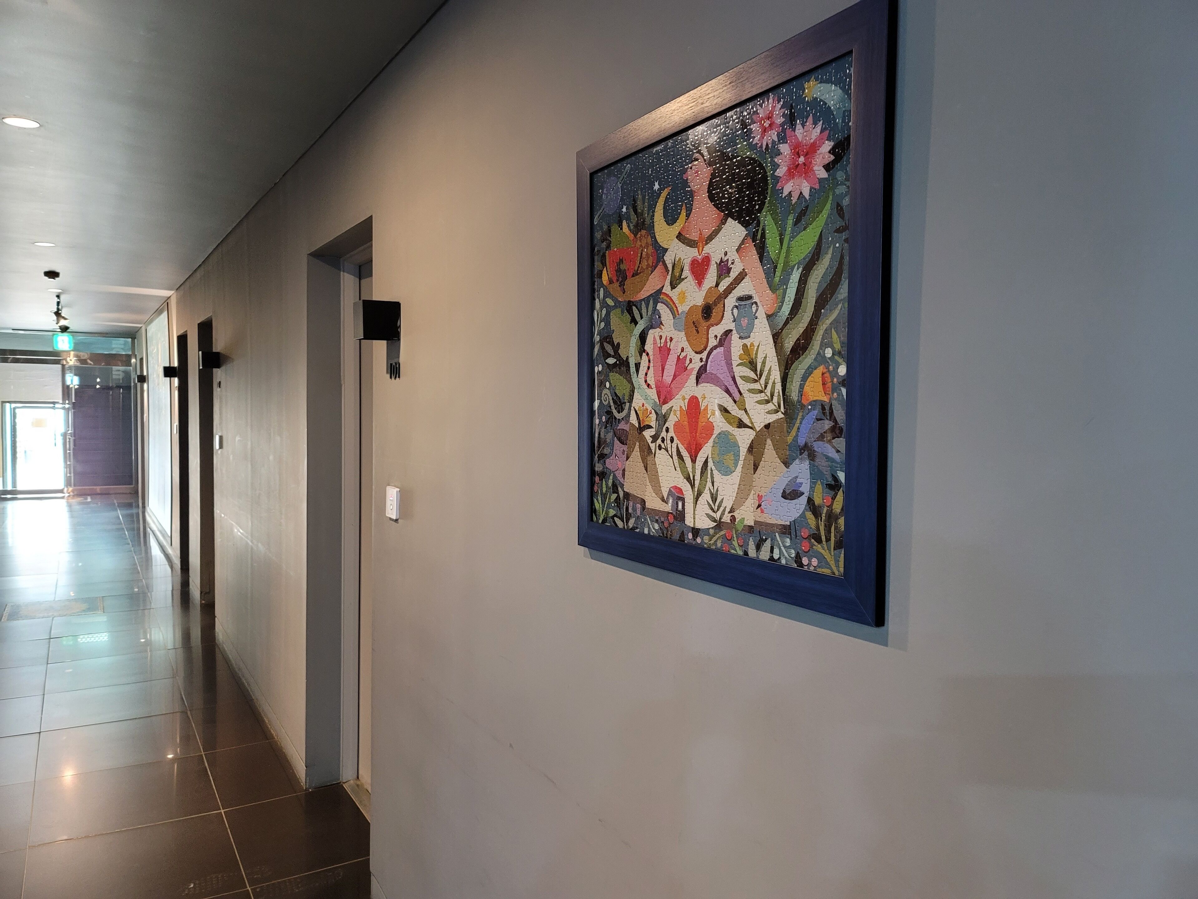 Een gang met een bloemig kunstwerk in een lijst aan de muur, een deur en een lichtschakelaar.