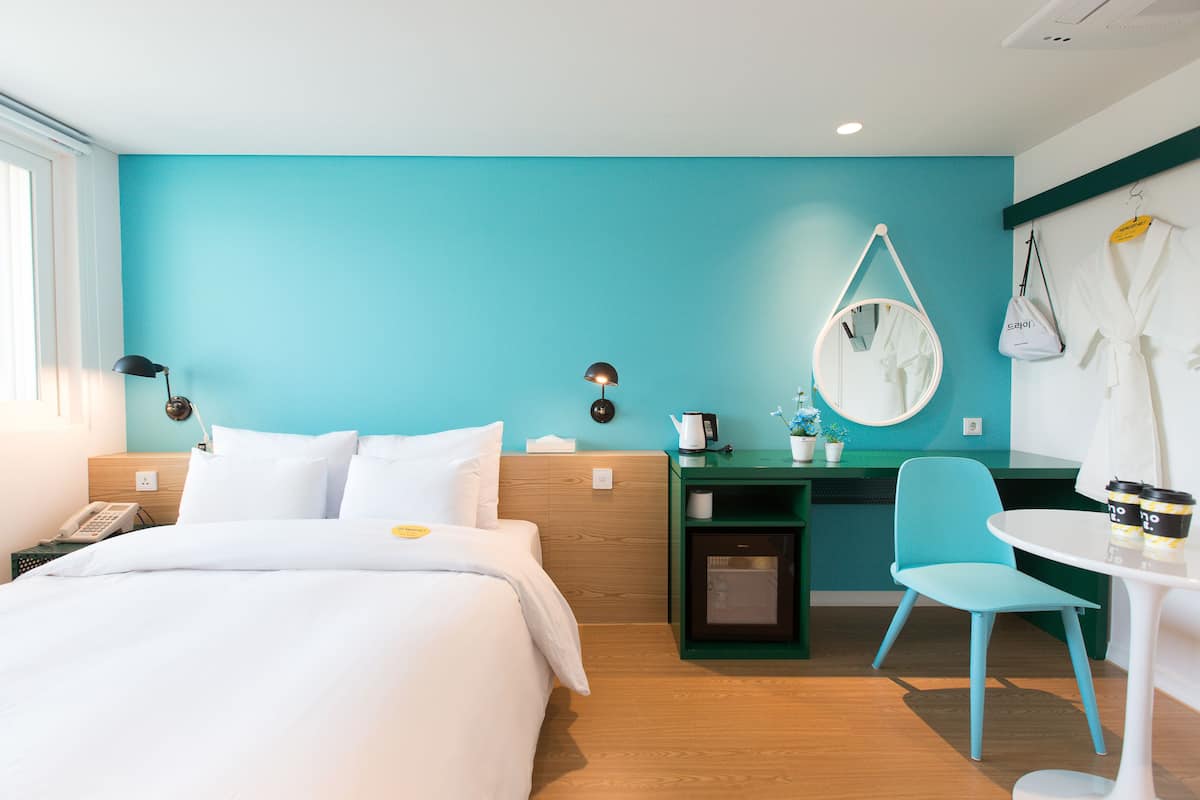 A hotel room with a bed, a desk, a chair, a mirror, and a table with a coffee cup.