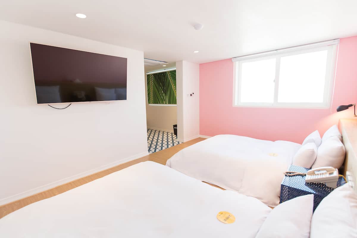 A hotel room with a flat-screen TV mounted on the wall, two beds with white bedding, a window with a view, and a small table with a lamp.