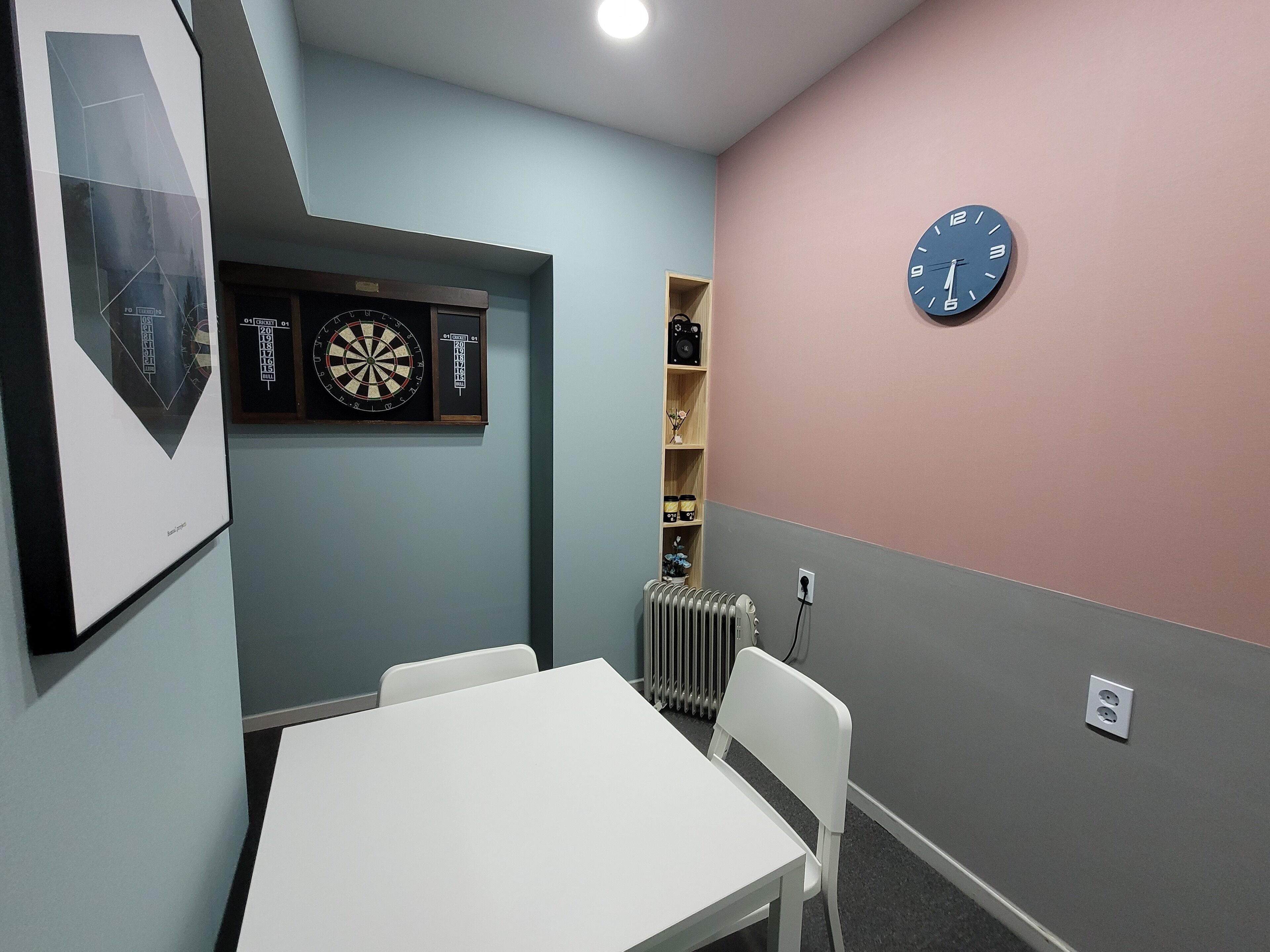 A room with a dartboard, a clock, a radiator, and a white table.