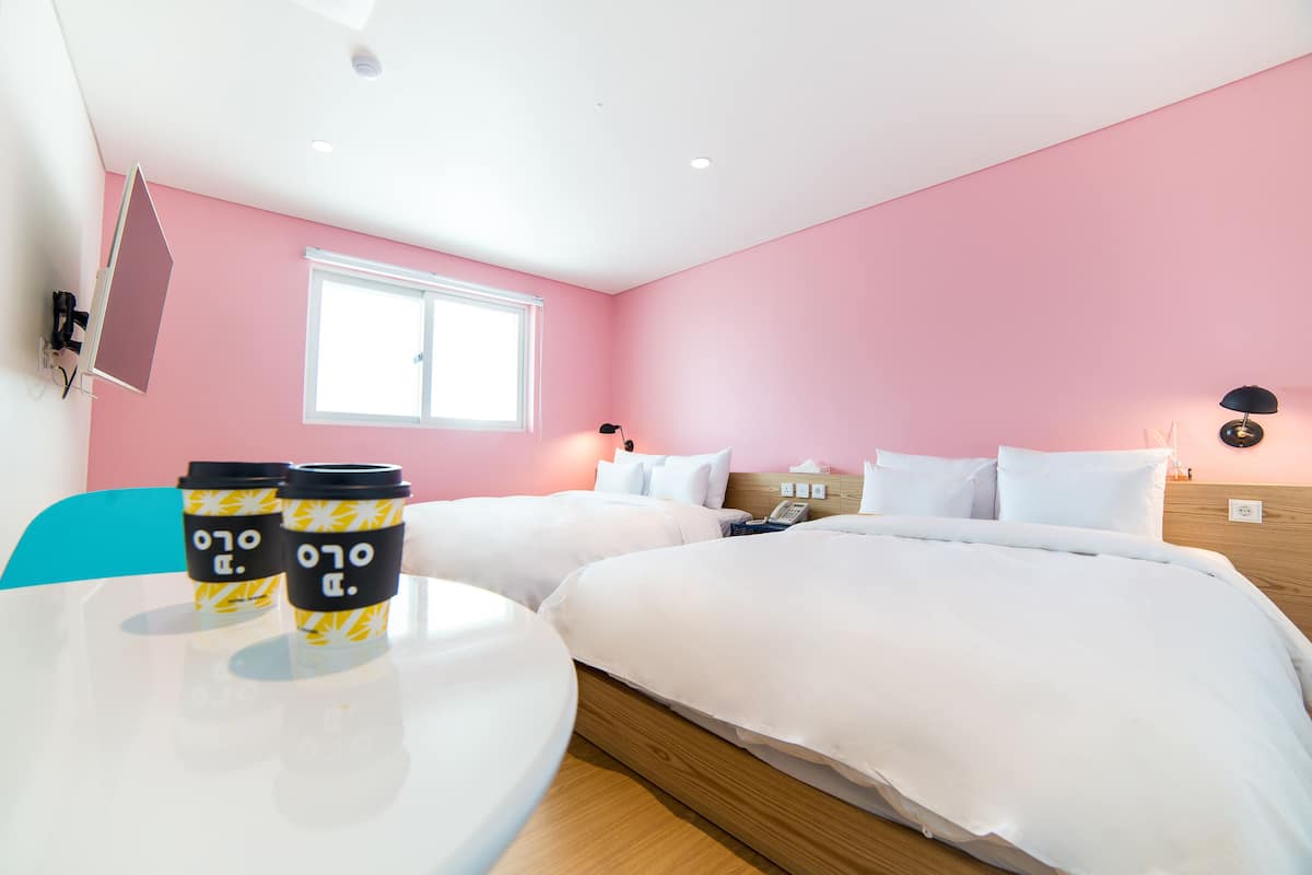 A hotel room with two beds, a window, a table with coffee cups, and a wall-mounted TV.